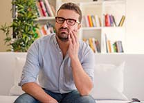 man sitting on his couch and holding his cheek in pain